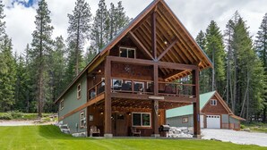 A two-story log cabin with a balcony, surrounded by a forest. - River's Bend Lodge- Hot Tub, Fire Pit, Games & Ski (Leavenworth)