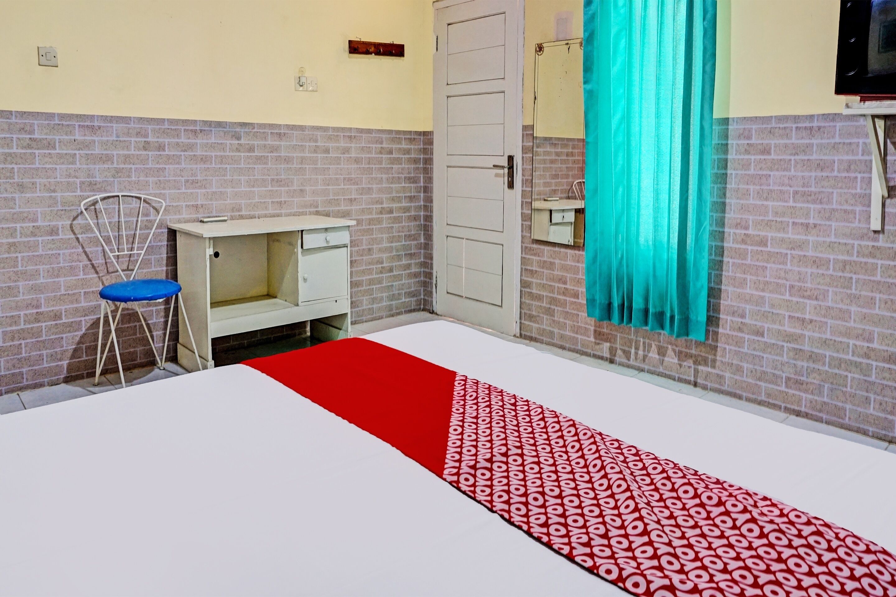 A hotel room with a white bed, a red and white patterned runner, a blue chair, a white cabinet, and a door with a mirror.