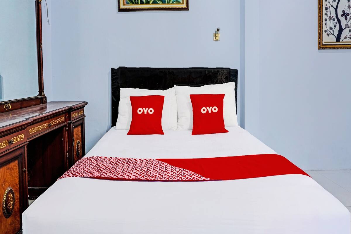 A hotel room with a bed, two red pillows with the word "OYO" on them, a wooden nightstand, and a mirror.