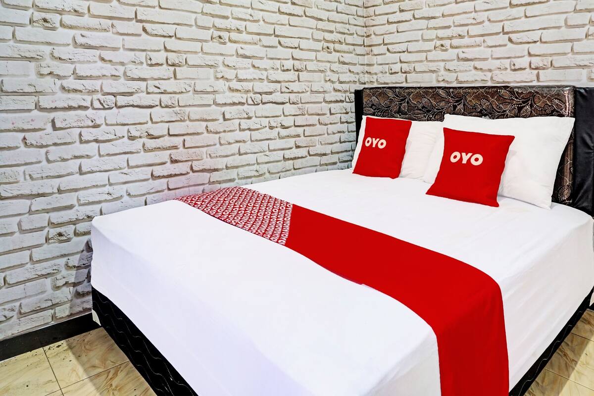 A neatly made bed with white and red bedding, two red pillows with the word "OYO" on them, and a patterned throw.