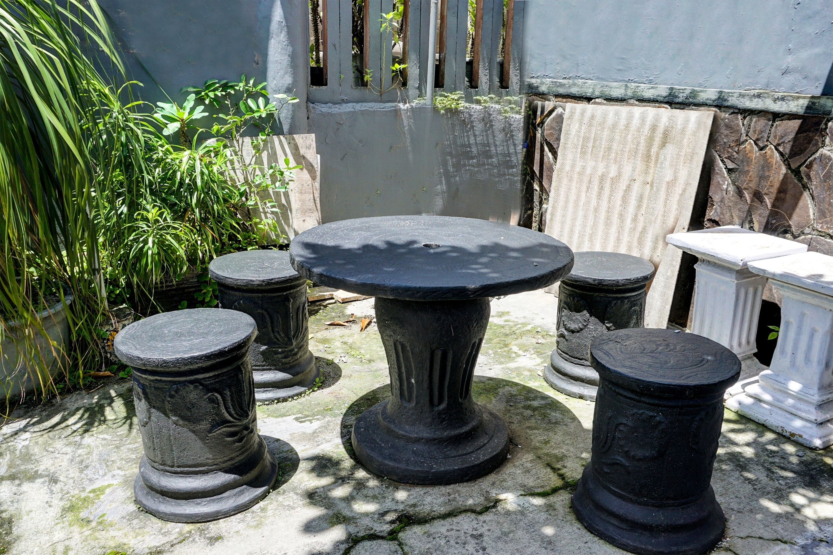 Une table en pierre, soutenue par quatre piédestaux assortis, se trouve dans une cour agrémentée de plantes et bordée d’un mur en pierre.