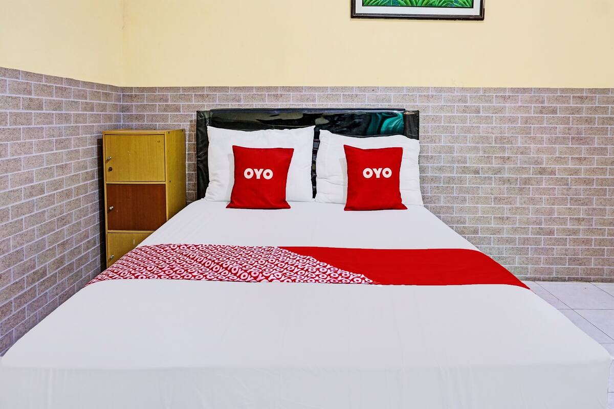A bed with a white and red bedspread, two red pillows with "OYO" text, a wooden wardrobe, and a brick-patterned wall.