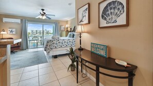 A hotel room with a bed, a desk, a chair, a potted plant, and a balcony with a view of the city. - Enchanted Inn Suite #5: Waterfront, Pool, Fishing Dock, Boat Docks & Tiki Bar! (Clearwater)