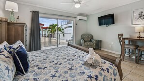 Ein Hotelzimmer mit Bett, Sessel, Tisch, Fernseher und einem Balkon mit Blick auf Palmen.
