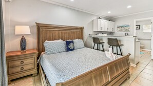 Ein Schlafzimmer mit einem hölzernen Bett, Nachttisch, Lampe und einer kleinen Küchenzeile mit Stuhl und Barhocker.