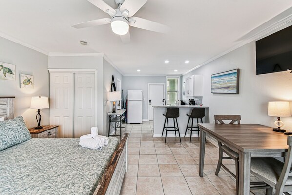 A hotel room with a bed, a desk, a chair, a table, a ceiling fan, and a kitchen area.