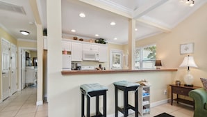 A kitchen with white cabinets, a microwave, and a coffee maker. There are two bar stools and a small table with a lamp. - Beachside Cottage 22 Steps to Beach | Beach Gear Included! (Clearwater)