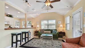 A spacious living area with a green sofa, a red armchair, and a white coffee table. There is a kitchen area with white cabinets and a bar counter. The room has a ceiling fan and recessed lighting. - Beachside Cottage 22 Steps to Beach | Beach Gear Included! (Clearwater)
