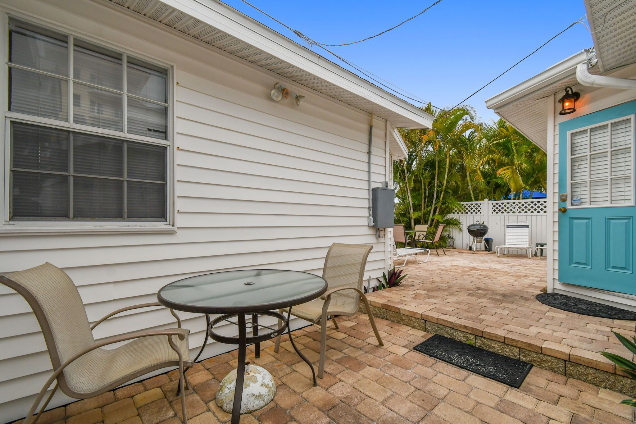 A patio with a table and chairs, a blue door, and a white fence with palm trees in the background.