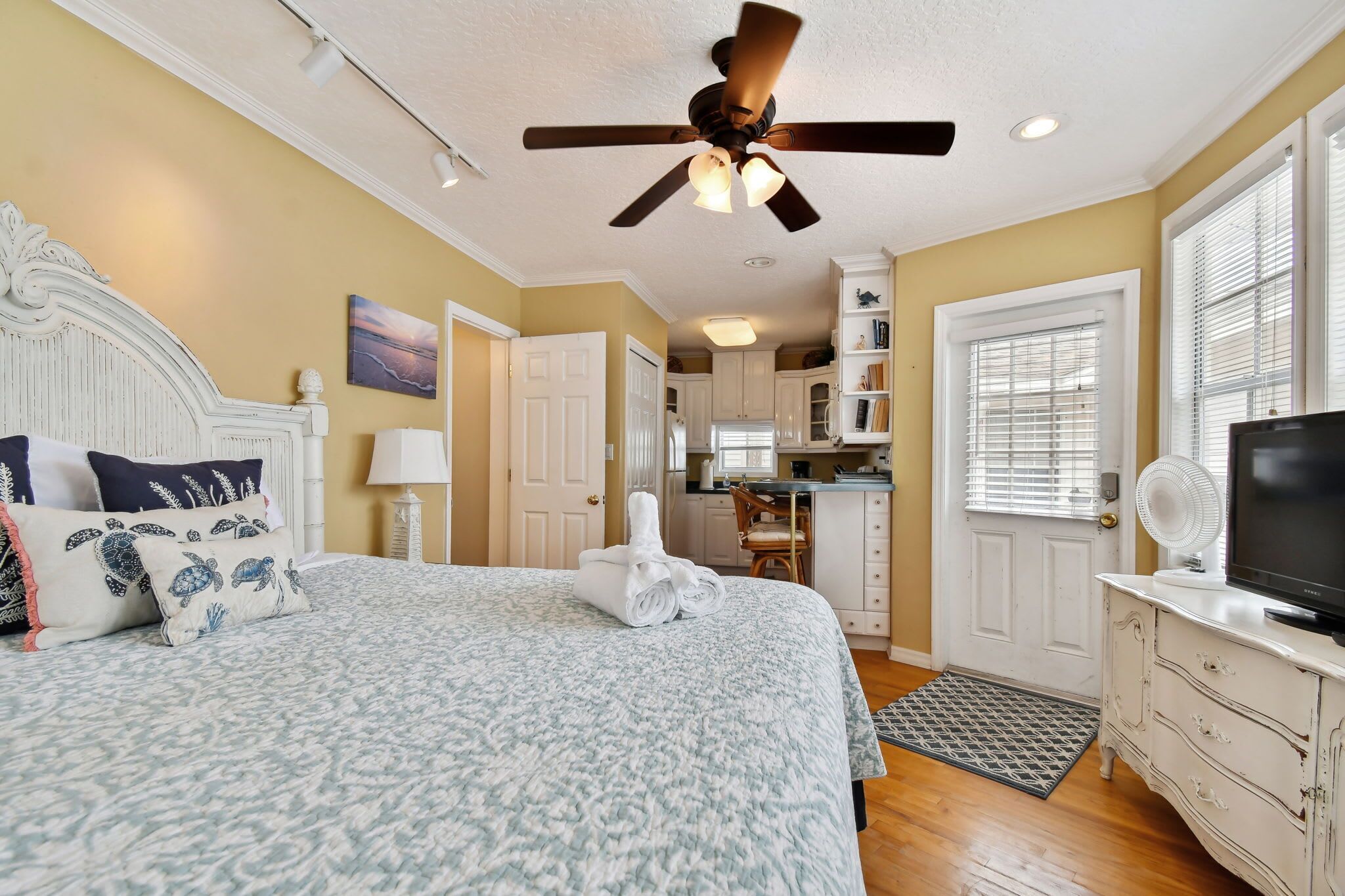 A bedroom with a bed, a ceiling fan, a desk, a chair, a television, and a kitchen area in the background.