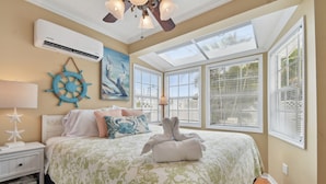 A bedroom with a large bed, a wall-mounted air conditioner, a ceiling fan, and a view through the skylight.