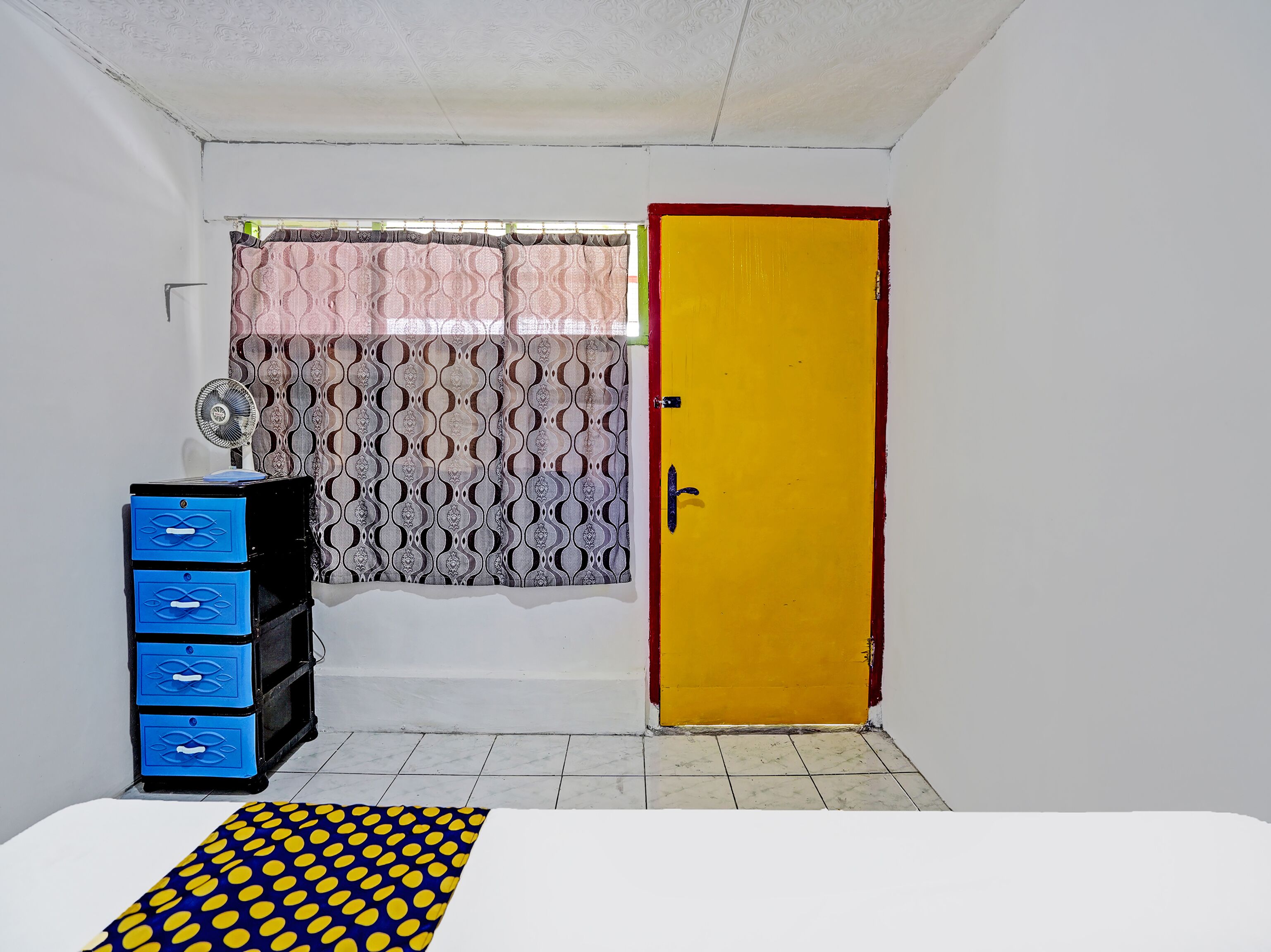 Una habitación pequeña y funcional con una cama, un mueble azul, una puerta amarilla, cortinas estampadas y suelo de baldosas.