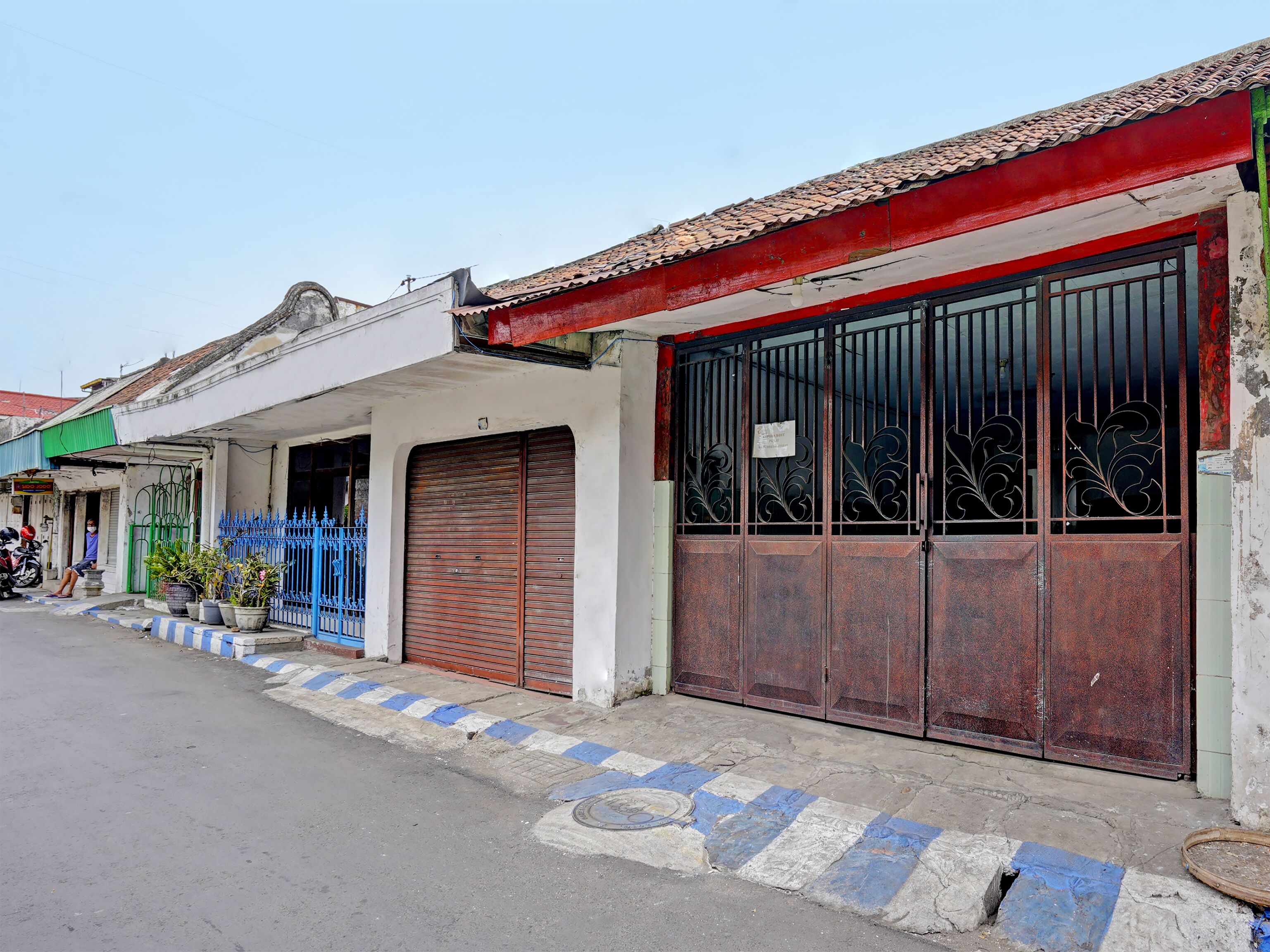 A street view with a building featuring a closed metal gate, a blue fence, and potted plants.