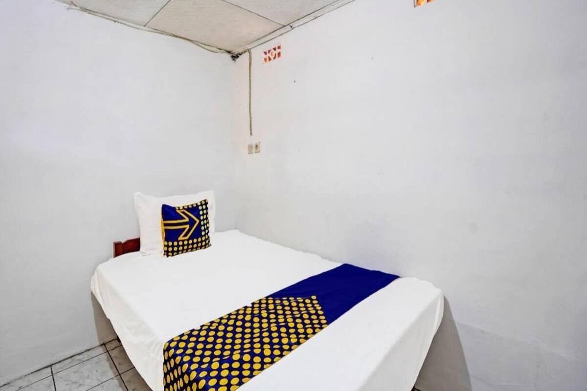 A small, single-bed room with a white ceiling, a visible electrical outlet, and a wall-mounted light switch.