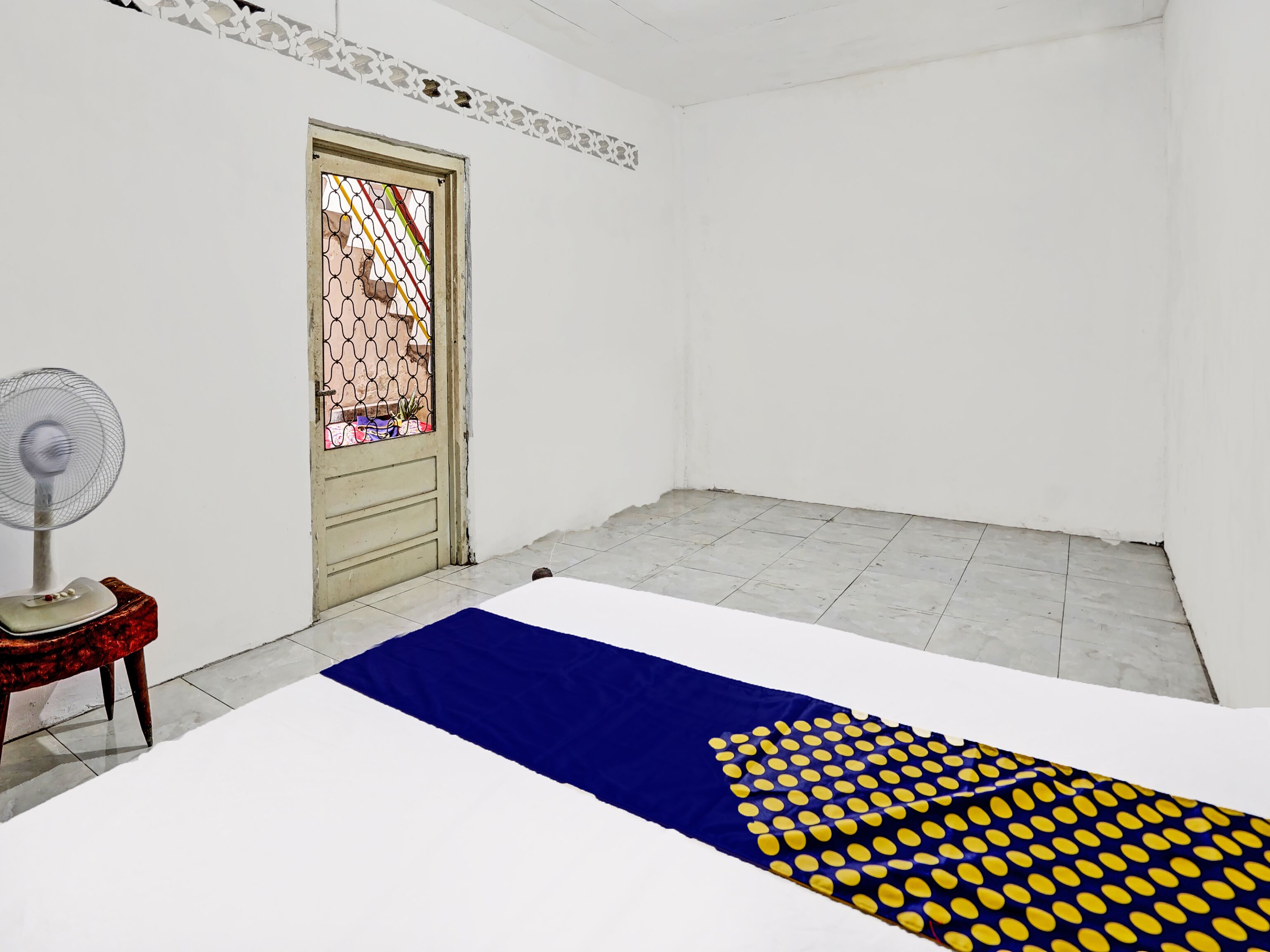 Una habitación con cama, ventilador de pie, puerta con celosía decorativa y suelo de baldosas.