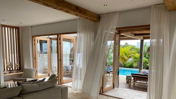 A spacious living room with a beige sofa, a glass door leading to a pool area, and sheer curtains. - Villa Bamboa – Ibiza-Styled Tropical Escape Near Jan Thiel Beach (Jan Thiel)