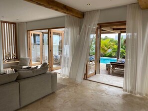 Amplia sala de estar con un sofá color beige, una puerta de cristal que da a una piscina y cortinas translúcidas.