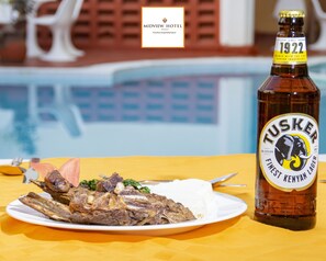Une bouteille de bière Tusker et une assiette de nourriture sur une nappe jaune, près d’une piscine.