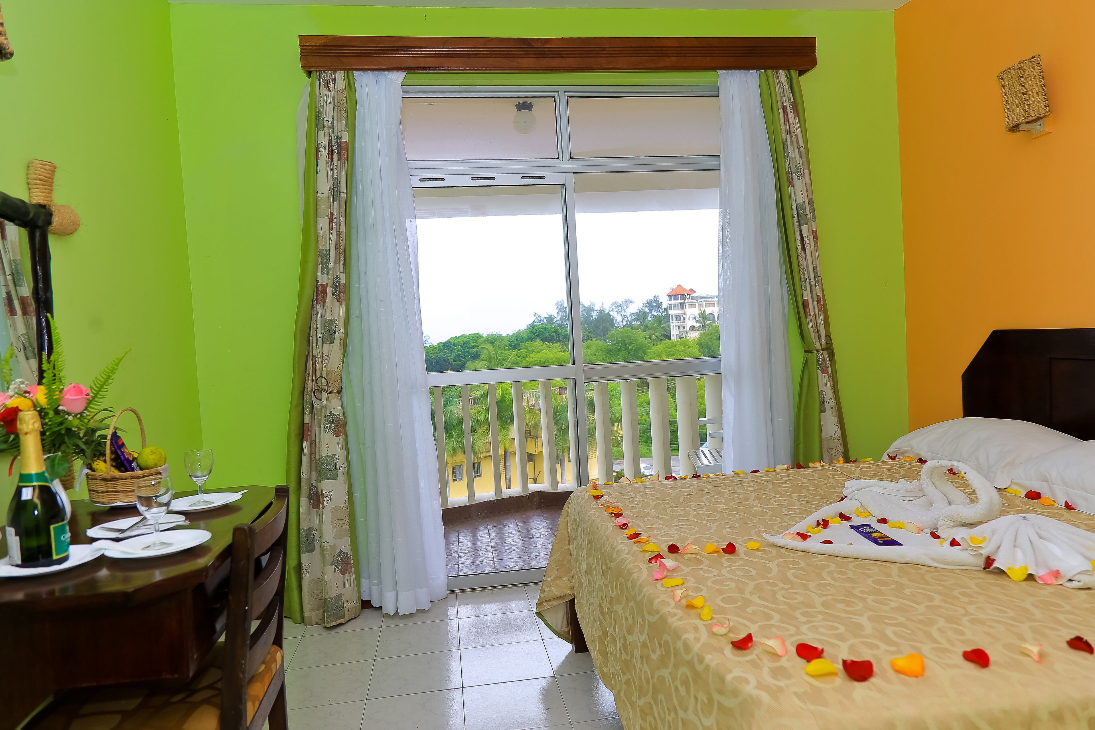 A hotel room with a balcony, a bed with rose petals, a dining table with a bottle and glasses, and a view of greenery and a building outside.