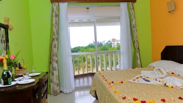 A hotel room with a balcony, a bed with rose petals, a dining table with a bottle and glasses, and a view of greenery and a building outside.