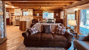A cozy cabin interior with a wooden ceiling, a stone fireplace, a brown leather sofa with plaid cushions, and a kitchen area in the background.