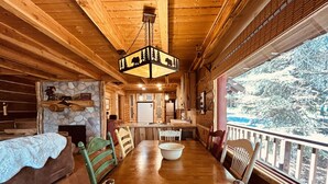 A cozy cabin interior with a wooden dining table, chairs, a fireplace, and a large window overlooking a snowy landscape.