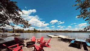 A lakeside setting with red Adirondack chairs, a fire pit, and a table under a clear sky. - Lakefront with private dock & fire pit - Bunk House Cabin (Island Park)