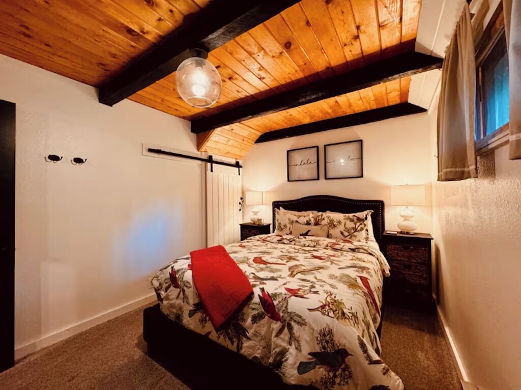 A bedroom with a wooden ceiling, a bed with a floral bedspread, a red throw pillow, and two framed pictures on the wall.