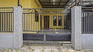 A gated entrance with a metal gate and decorative pillars, leading to a building with a yellow facade and a covered area with a metal roof.