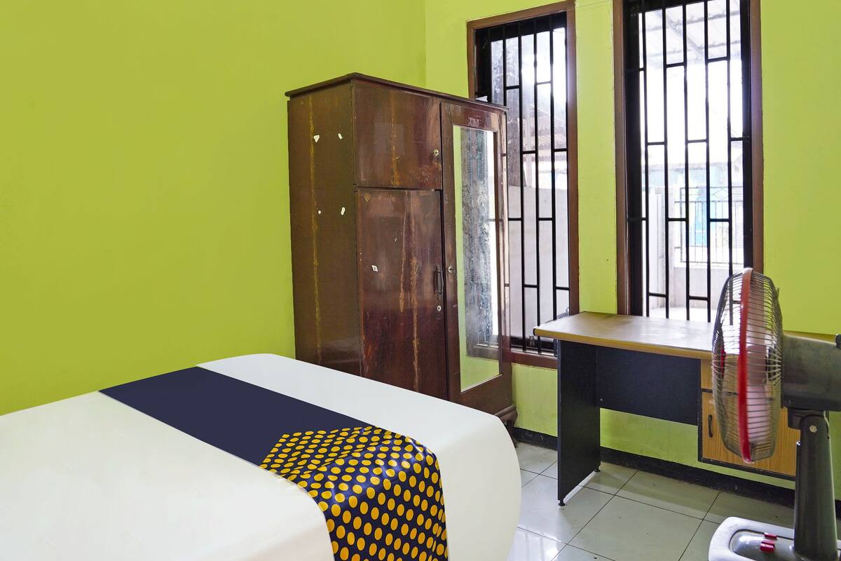 A room with a bed, a wooden wardrobe, a desk with a chair, and a standing fan.
