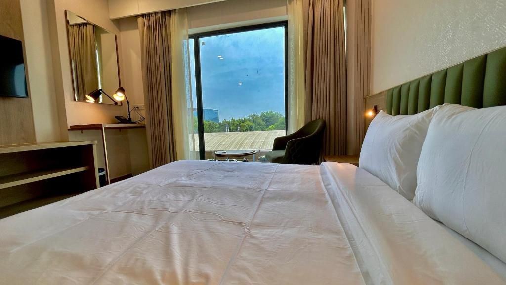 A hotel room with a large bed, a desk with a lamp, a chair, and a view of the outdoors through a large window.