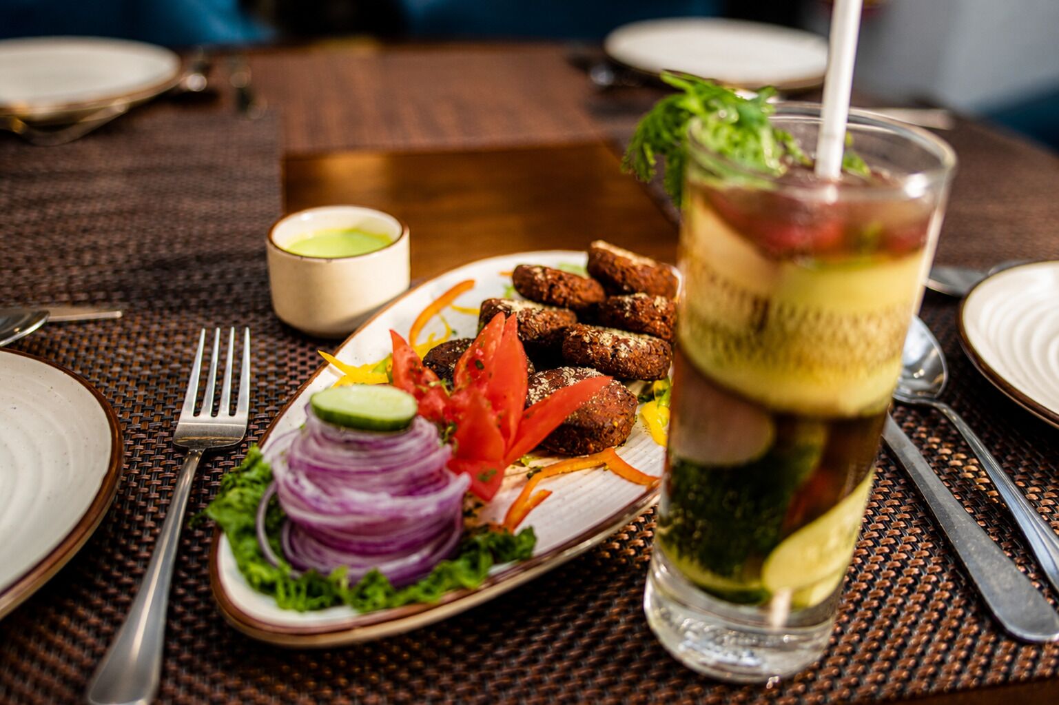 A dining table with a plate of falafel, a glass of iced tea, and a small bowl of sauce.