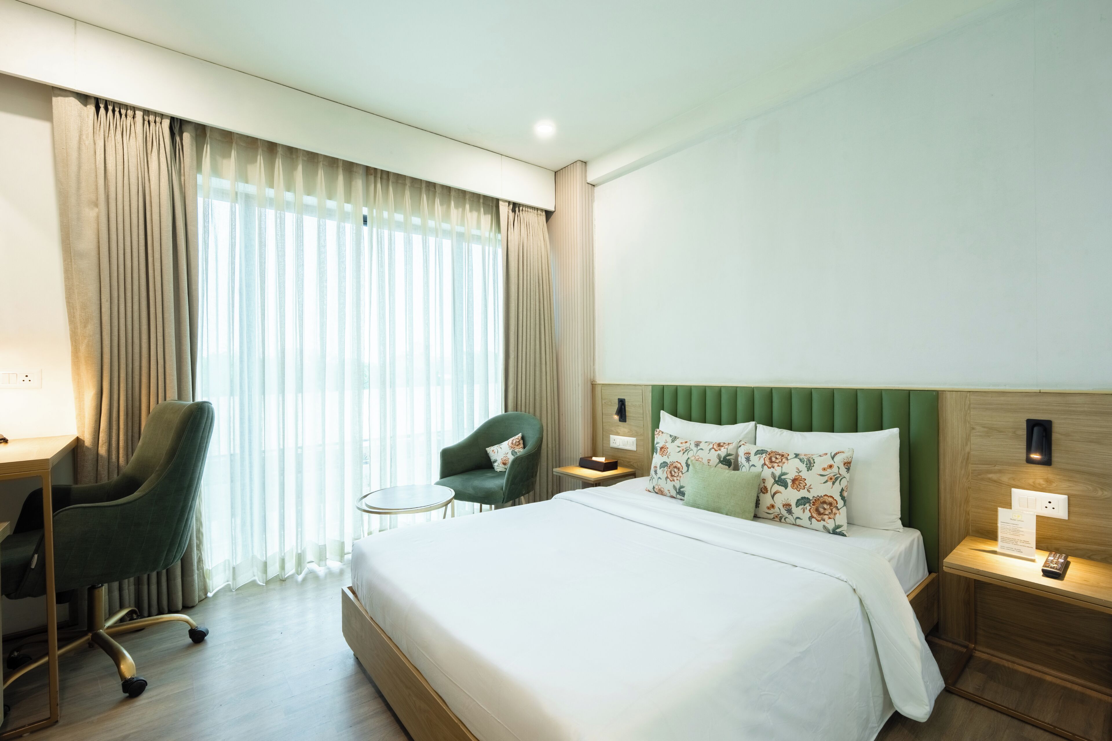 A hotel room with a bed, a desk, a chair, a small table, and a nightstand with a lamp.