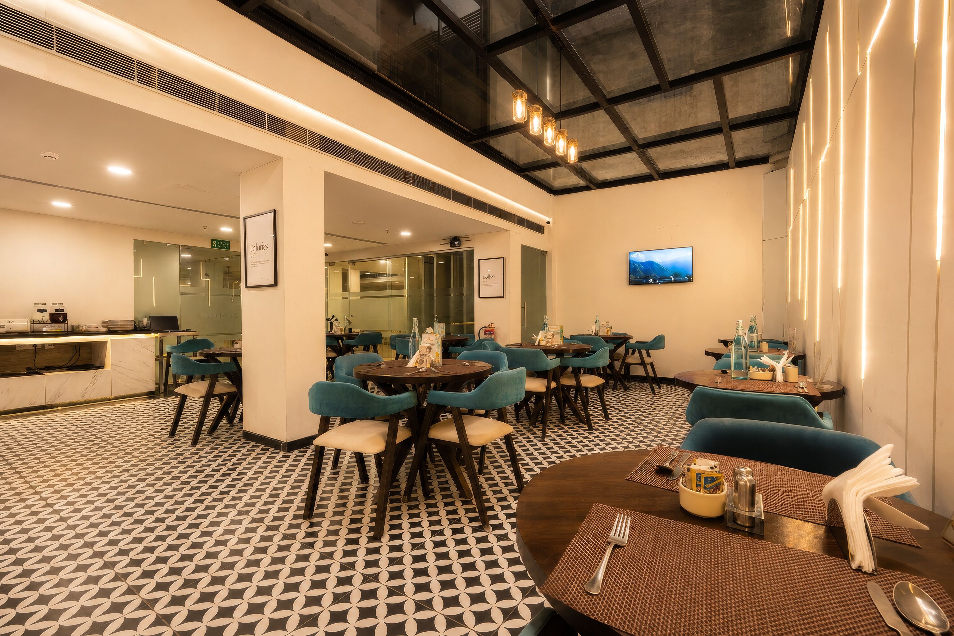 A modern hotel lobby with a geometric patterned floor, multiple seating areas with blue chairs, a bar area, and a television mounted on the wall.