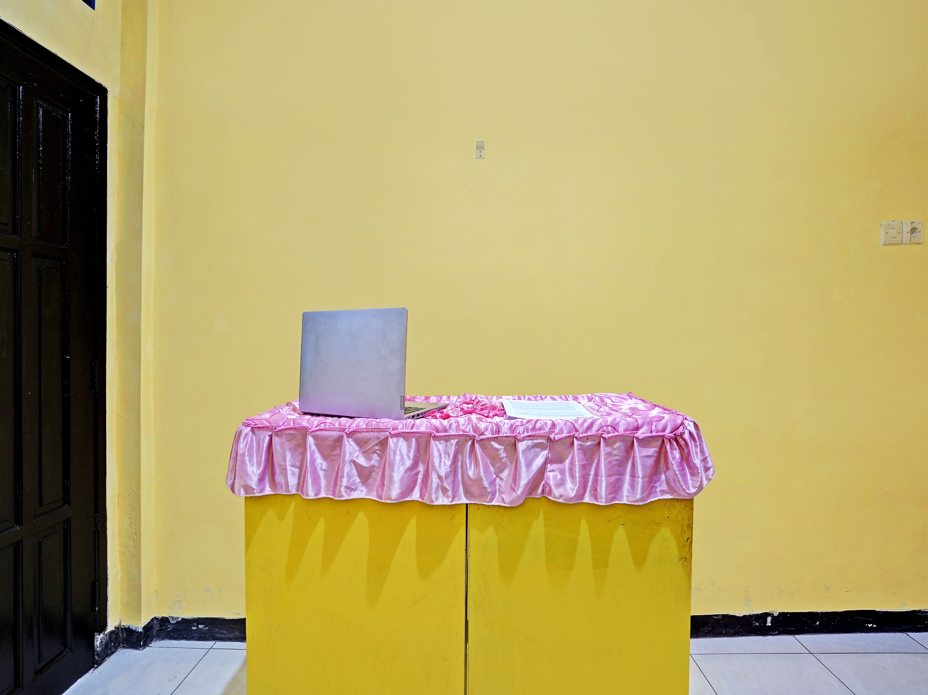 Een gele tafel met een laptop en een roze kleed, voor een gele muur.