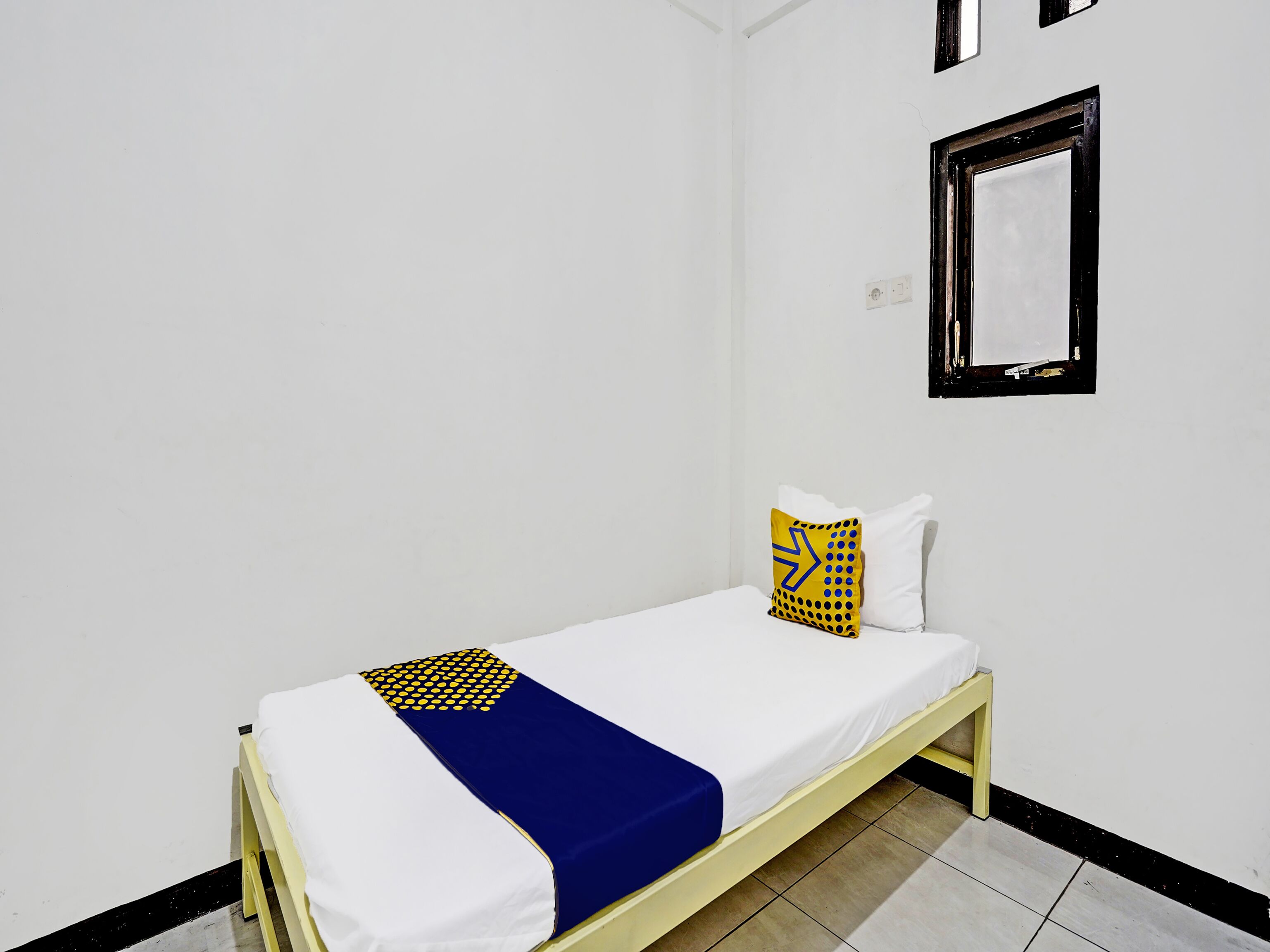 A small, single-bed room with white walls, a black-framed window, and a yellow and blue bedspread.