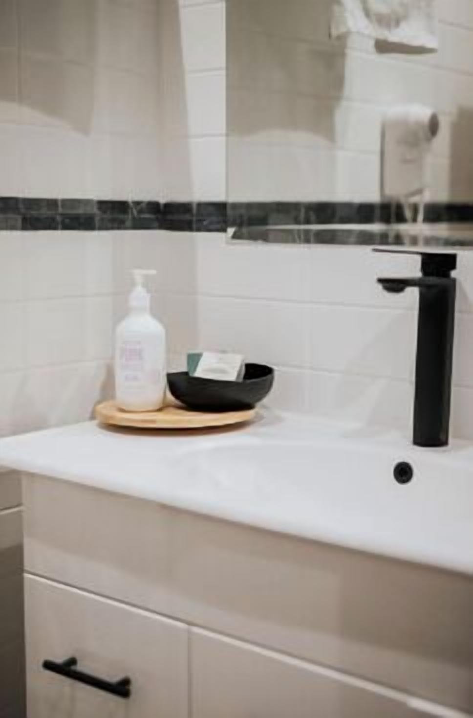 A bathroom with a white countertop, a black faucet, a soap dispenser, and a black bowl on a wooden tray.