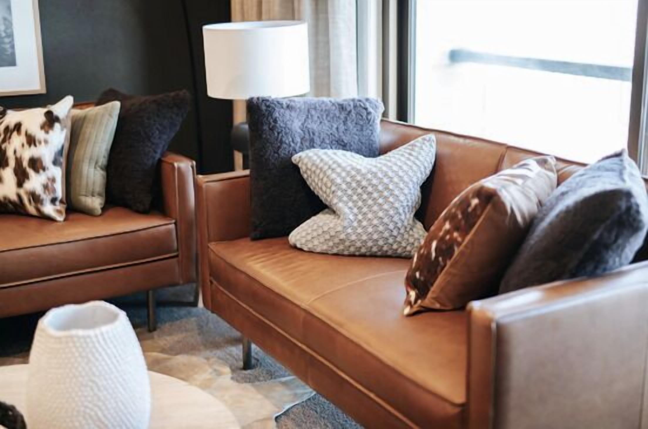A modern living room with a brown leather sofa, patterned cushions, a white vase, and a lamp.