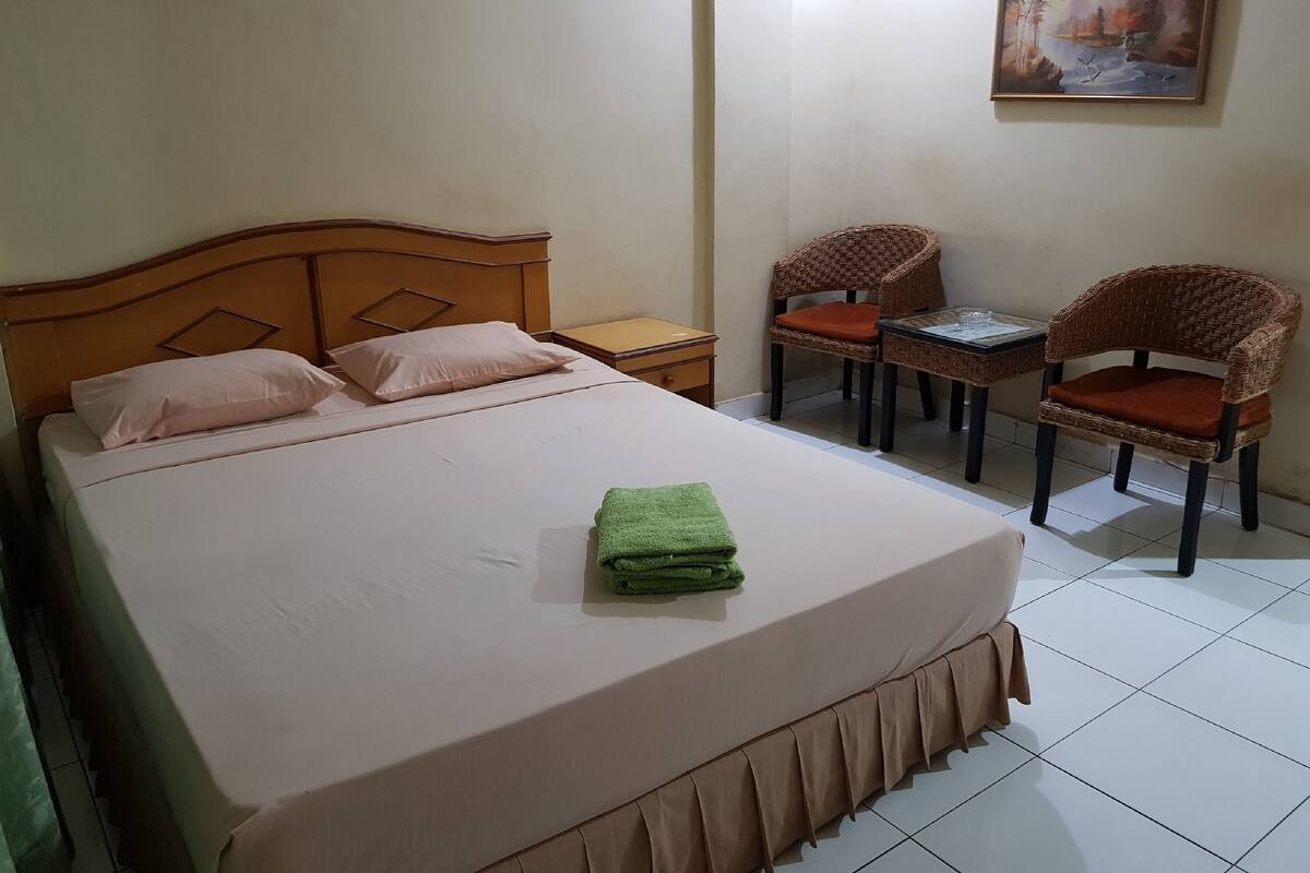 Un lit bien fait, avec une serviette verte, deux chaises en osier, une petite table et un tableau accroché au mur.