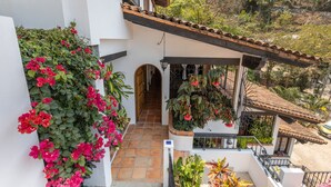 Un edificio blanco con techo de tejas, un balcón con plantas colgantes y vista a un paisaje montañoso.