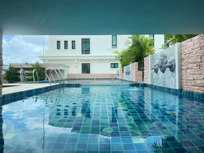 Una piscina amb rajoles blaves cristal·lines, una paret de pedra amb decoracions de caps de lleó i un edifici blanc al fons.
