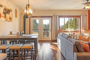 A spacious living area with a sofa, a dining table with stools, a chandelier, and large windows offering a view of the outdoors.