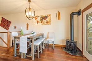 Un coin repas avec une table en bois, des bancs, un lustre, une œuvre d’art représentant une carte du monde, un poêle rustique et une fenêtre offrant une vue agréable.