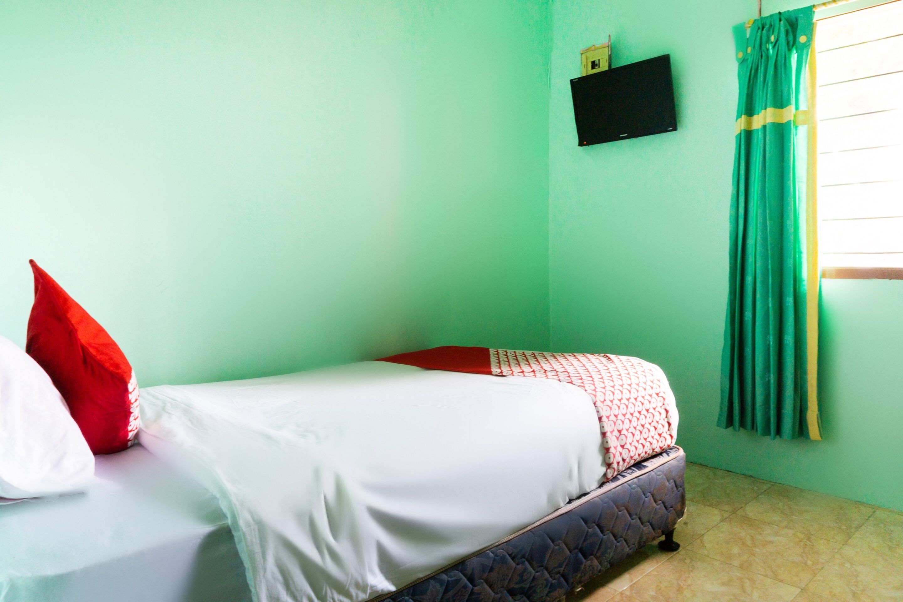 Una cama individual con ropa de cama blanca y roja, un televisor pequeño en la pared y una ventana con cortinas verdes.