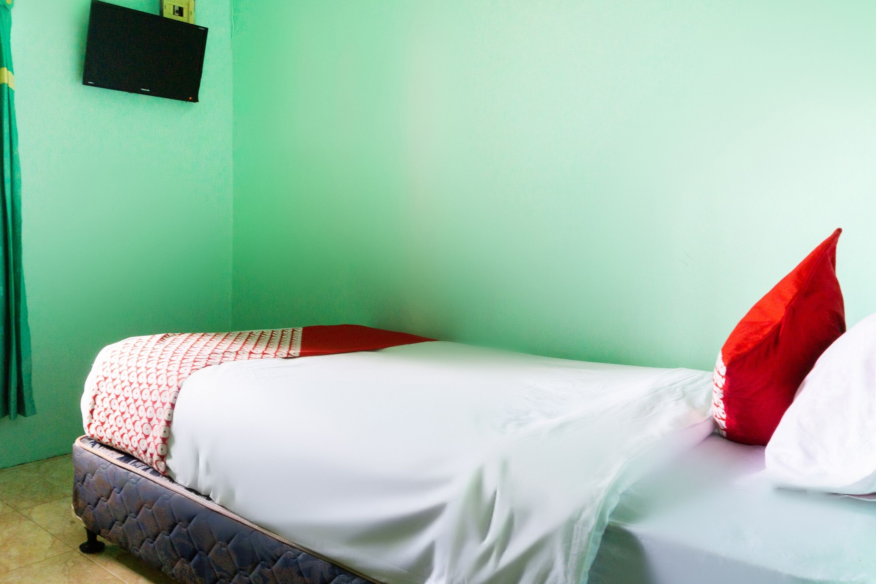 A single bed with a quilted headboard, a flat-screen TV mounted on the wall, and a green curtain.
