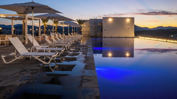 A poolside area with lounge chairs and umbrellas, a clear view of the mountains, and a calm water surface reflecting lights.