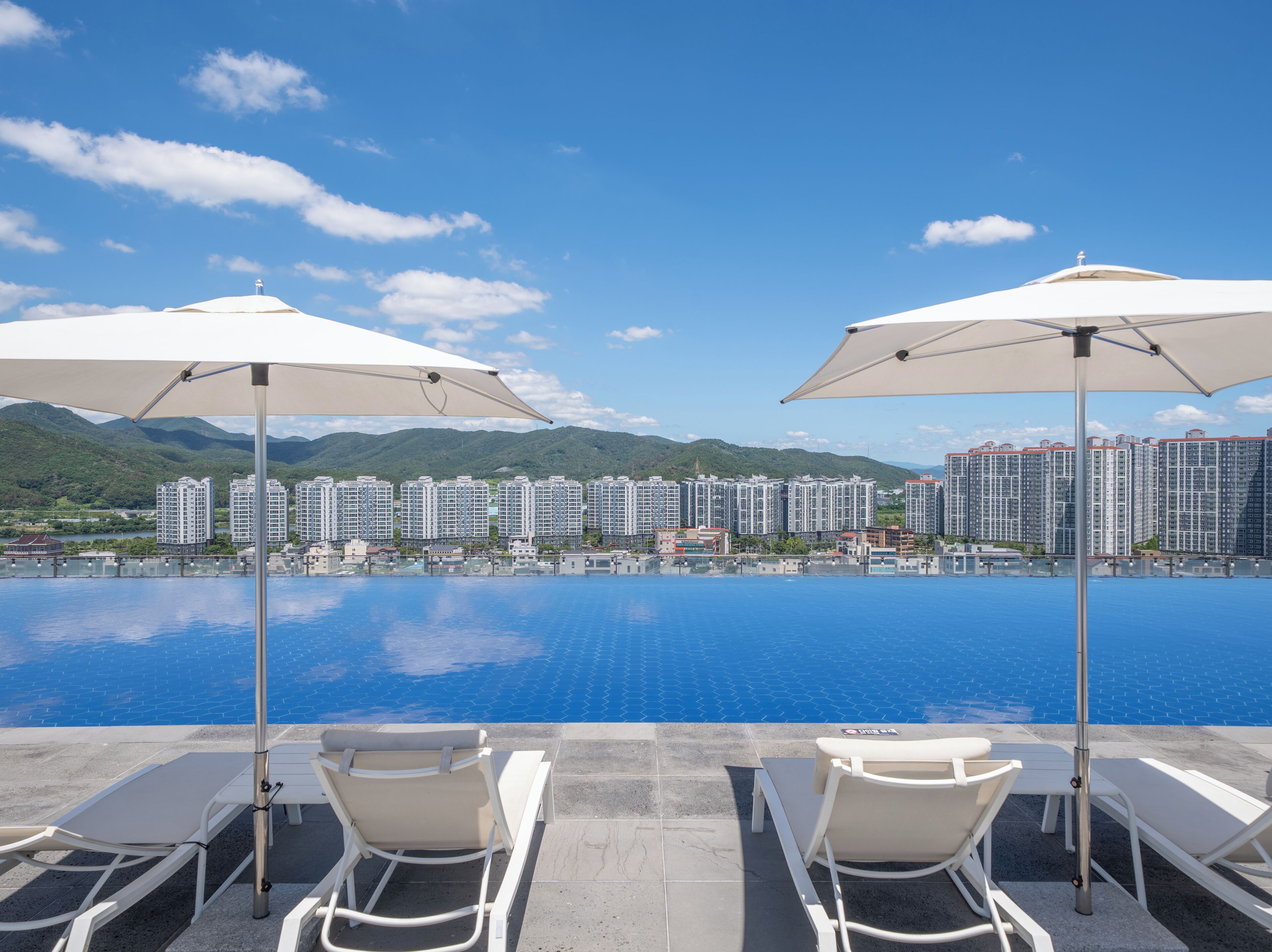 A poolside area with lounge chairs and umbrellas, overlooking a large body of water and a cityscape with high-rise buildings.