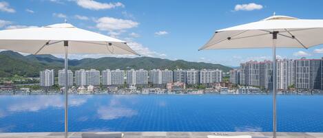 A poolside area with lounge chairs and umbrellas, overlooking a large body of water and a cityscape with high-rise buildings.