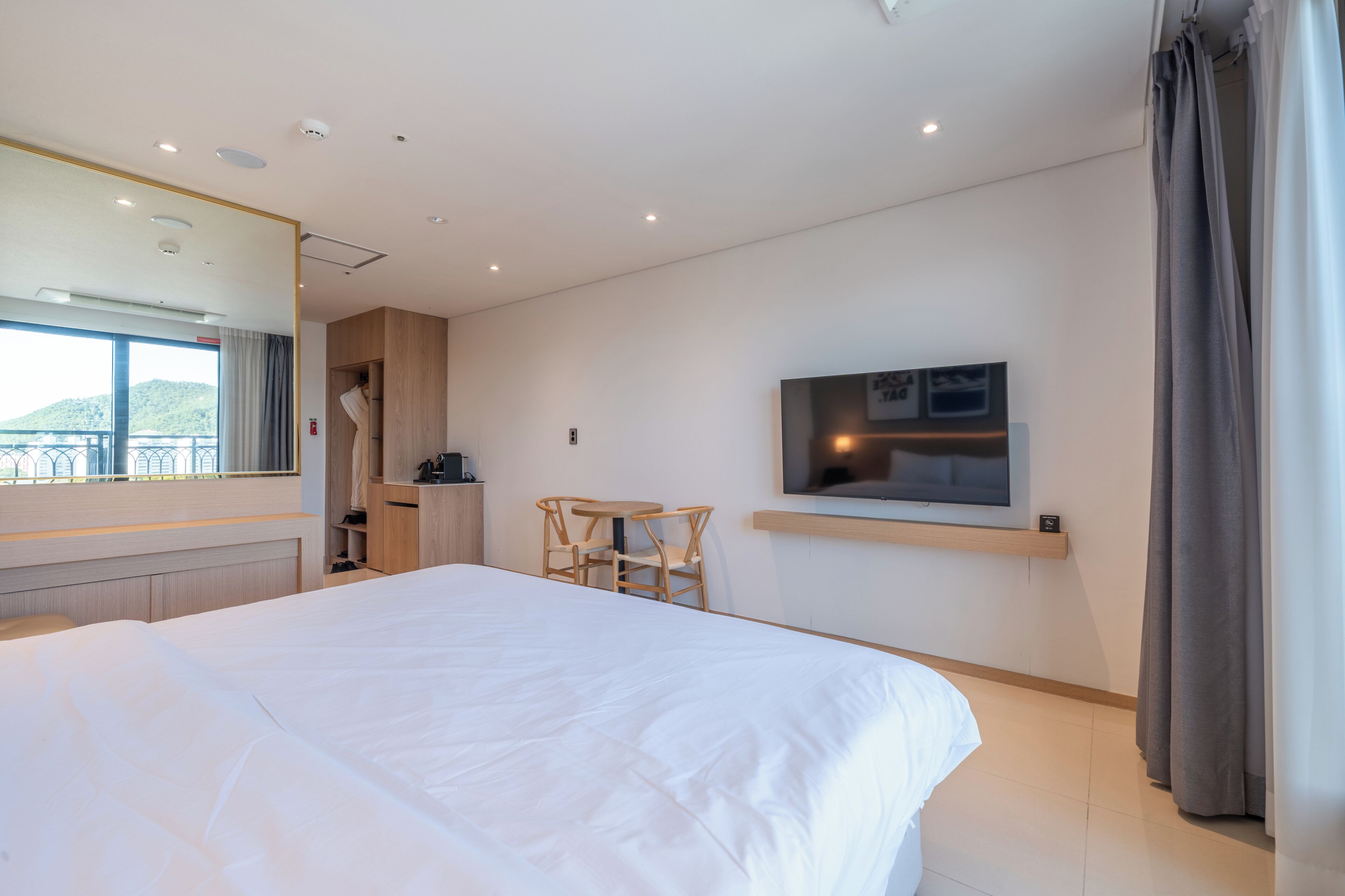 A hotel room with a large bed, a TV mounted on the wall, a small desk with a chair, and a window with curtains.