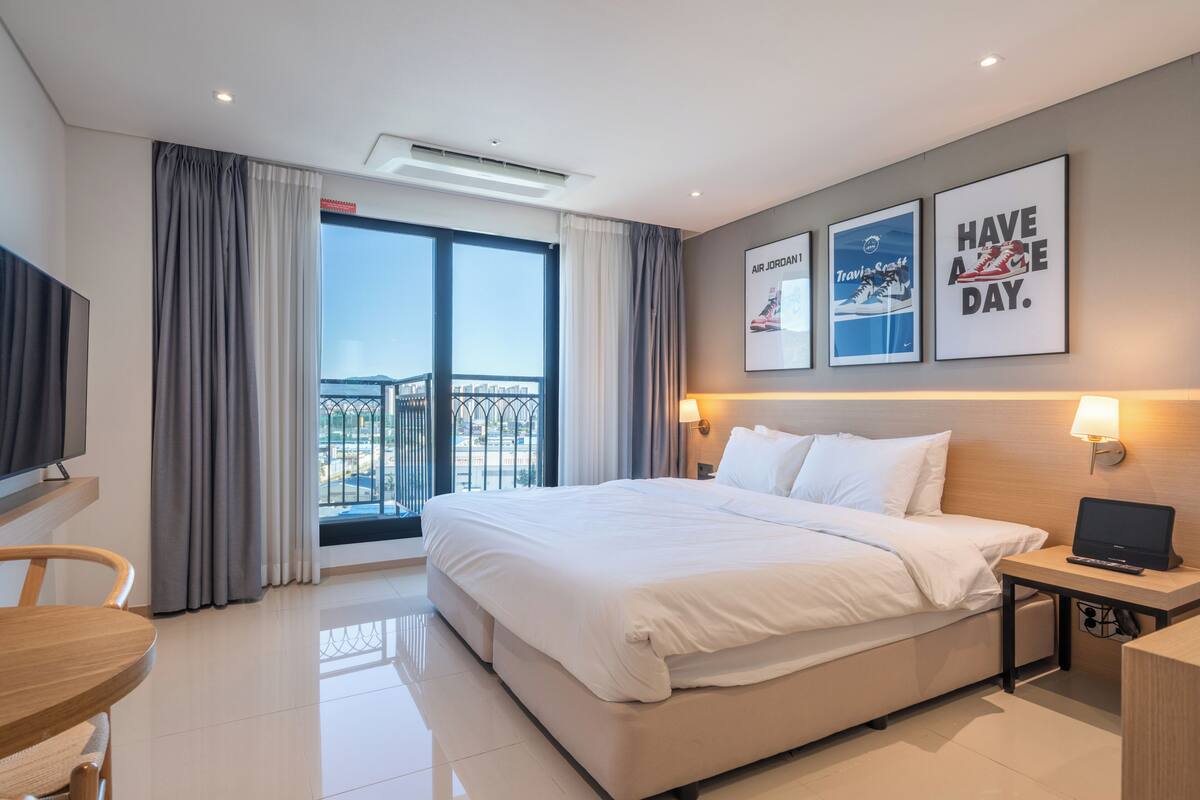 Een hotelkamer met een groot bed, een bureau met een laptop, een televisie en een balkon met gordijnen.